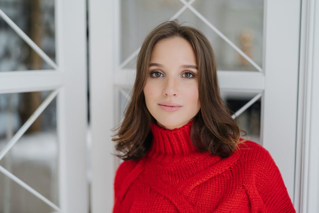 Calm beautiful young European woman wears warm red sweater, has make up and healthy soft skin